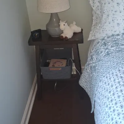Sale Photo Thumbnail #55: Dark wood nightstand holding a grey ceramic table lamp with a beige fabric shade, plush llama toy, and alarm clock, shown next to a bed with blue and white floral bedding.