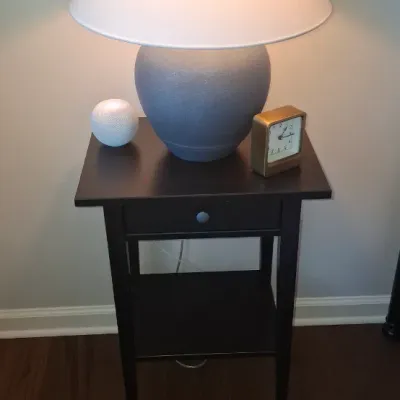 Sale Photo Thumbnail #38: Contemporary grey urn-style table lamp with a white drum shade, displayed on a dark brown wooden nightstand alongside a small brass table clock and a HomePod Mini.