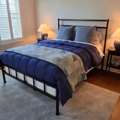 Sale Photo Thumbnail #40: Black metal queen bed frame set up with a navy comforter, decorative pillows, and a patchwork throw quilt, flanked by matching nightstands with lamps.