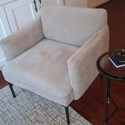 Sale Photo Thumbnail #3: Contemporary beige upholstered armchair featuring slim black metal legs, set alongside a small glass-top side table on an area rug.