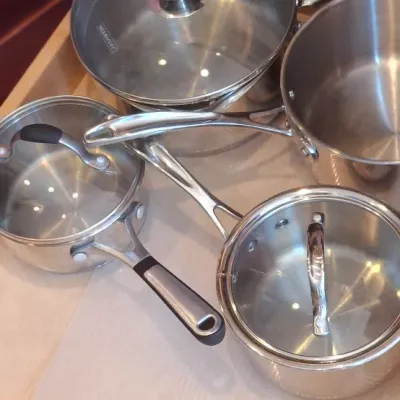 Sale Photo Thumbnail #79: Assorted stainless steel saucepans with glass lids stored in a pull-out wooden cabinet drawer, in used condition.
