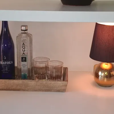 Sale Photo Thumbnail #20: Rustic wooden serving tray with Saratoga blue glass water bottles, liquor bottle, and drinking glasses, alongside a small modern spherical lamp and a picture frame.