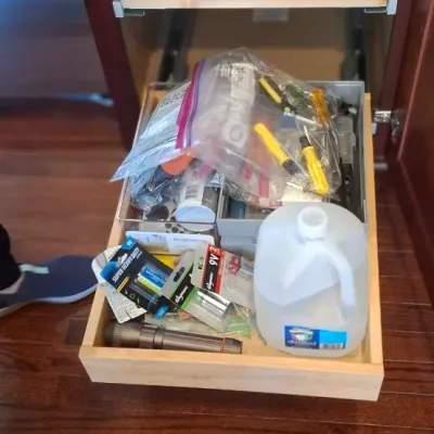 Sale Photo Thumbnail #74: Pull-out kitchen cabinet drawers containing various household supplies, including plastic wrap, small hand tools, batteries, a flashlight, and a jug of distilled water.
