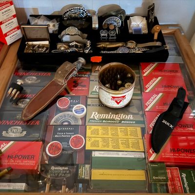 Sale Photo Thumbnail #124: Glass display case filled with vintage ammunition boxes, sheath knives, belt buckles, wristwatches, and small collectibles.