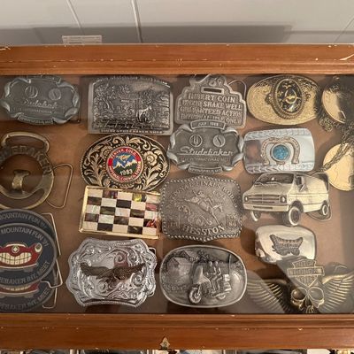 Sale Photo Thumbnail #120: Collection of vintage brass, silver-tone, and mother-of-pearl belt buckles displayed inside a wooden glass-top case, featuring Studebaker, rodeo, and novelty motifs.