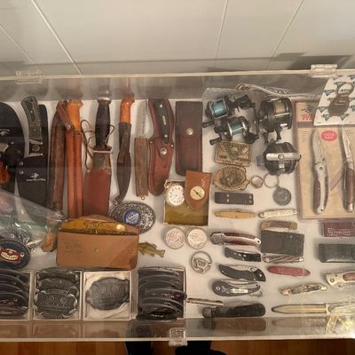Sale Photo Thumbnail #119: Vintage collectibles, including hunting knives with leather sheaths, fishing reels, pocketknives, belt buckles, and a pocket watch.
