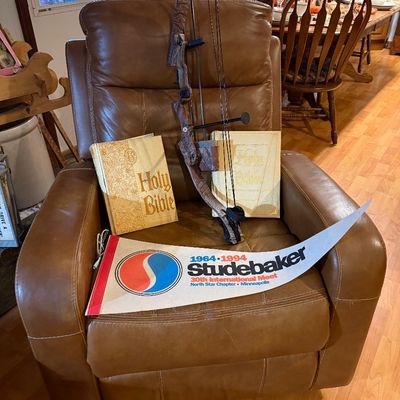 Sale Photo Thumbnail #110: Brown leather arm chair displaying a camouflage compound hunting bow, two hardcover Holy Bibles, and a 1994 Studebaker 30th International Meet felt pennant.