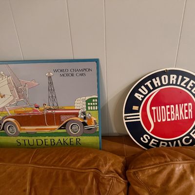 Sale Photo Thumbnail #111: Two vintage-style Studebaker advertising signs, including a rectangular poster featuring a classic car and airplane, circular round porcelain-style 'Authorized Service' emblem sign.