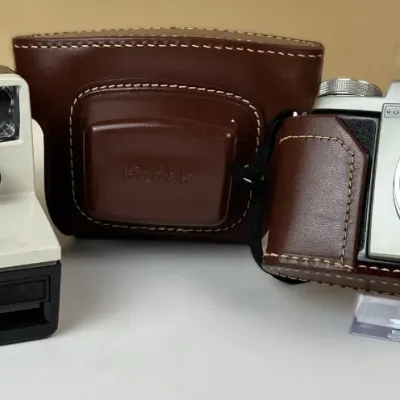 Sale Photo Thumbnail #142: Vintage Polaroid OneStep Land camera paired with a Kodak Pony IV 35mm camera and its brown leather case, circa 1950s-1970s.
