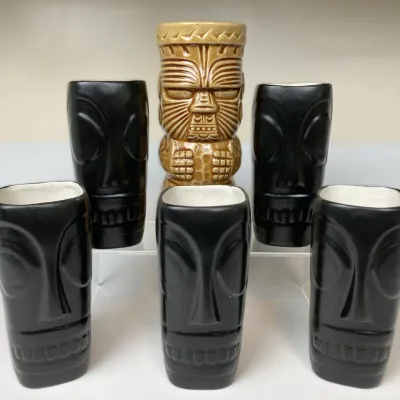 Sale Photo Thumbnail #46: Set of vintage ceramic tiki mugs, including five black Moai-style tumblers with white interiors and one taller carved tan tiki statue mug.