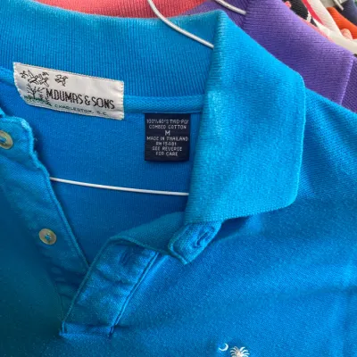 Sale Photo Thumbnail #162: Bright blue M. Dumas & Sons polo shirt featuring an embroidered palmetto tree and crescent moon logo, hanging alongside other colorful shirts.