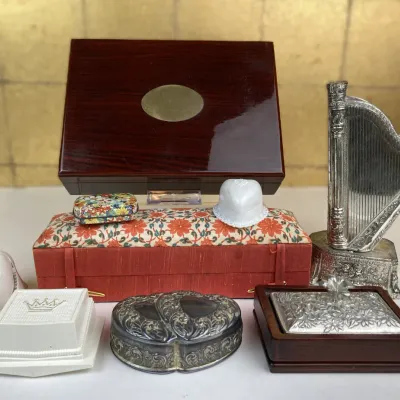 Sale Photo Thumbnail #243: Collection of decorative trinket boxes and small collectibles, including a polished wooden box with a brass medallion, a silver-tone mini harp, a floral silk fabric box, and various vintage ceramic and metal cases.