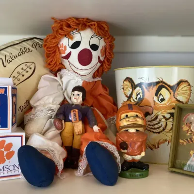 Sale Photo Thumbnail #139: Collection of vintage Clemson Tigers memorabilia including rag dolls, a wastebasket, figurines, framed art, and a plush football.