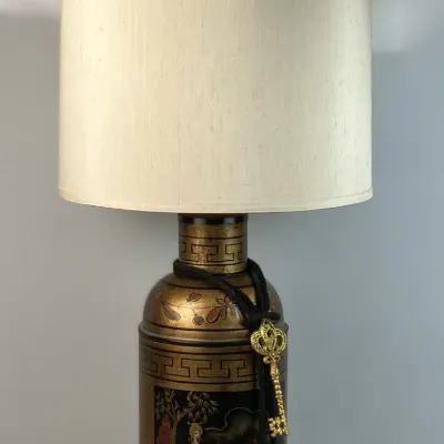Sale Photo Thumbnail #11: Vintage Asian-inspired tea canister table lamp featuring a hand-painted gilt and black Chinoiserie landscape design, black tassel with key ornament, and tall cream drum shade.