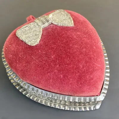 Sale Photo Thumbnail #237: Vintage heart-shaped trinket box featuring a red velvet top decorated with an embroidered silver bow applique and a fluted silver-tone metal base.
