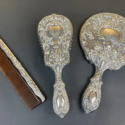 Sale Photo Thumbnail #240: Vintage three-piece sterling silver vanity set including a handheld mirror, hairbrush, and comb, featuring intricate Victorian-style repoussé floral scrollwork.