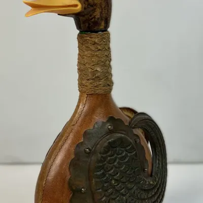Sale Photo Thumbnail #65: Vintage duck-head decanter encased in stitched brown leather with embossed metal wing accents and a yellow bill stopper.