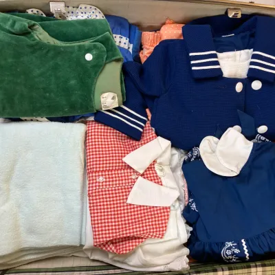 Sale Photo Thumbnail #246: Open vintage hardshell suitcase packed with retro children's clothing, including sailor-style dresses, a red gingham shirt, and a green velvet garment.