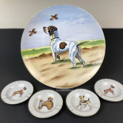 Sale Photo Thumbnail #223: Vintage hand-painted porcelain dog scene display plate featuring a hunting hound, accompanied by four matching small individual dog-themed ash trays.
