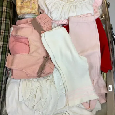 Sale Photo Thumbnail #247: Vintage plaid-lined hardshell suitcase packed with baby clothes, including pink corduroy overalls, knit tights, dresses, and a boxed pair of baby shoes.