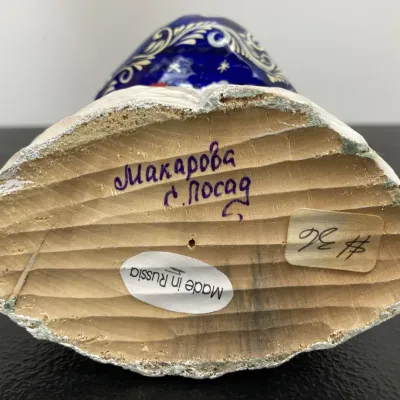 Sale Photo Thumbnail #210: Underside of a hand-carved Russian wooden figurine, showing a hand-written artist signature, "Made in Russia" sticker, and price tag.