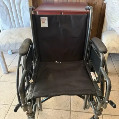 Folding manual wheelchair with a dark metal frame and black fabric seating, featuring padded armrests and front caster wheels.