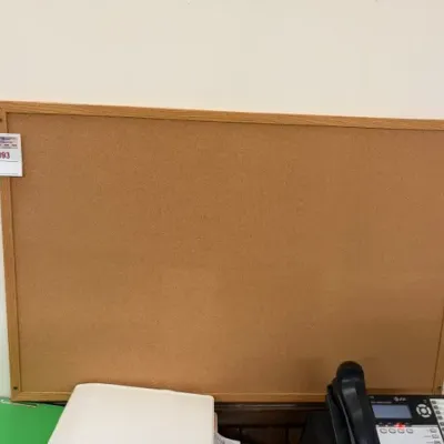 Large rectangular cork bulletin board framed in light oak wood, shown mounted on a white wall above office supplies.
