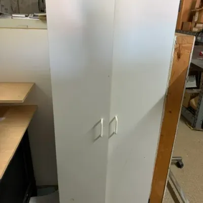 Tall white two-door storage cabinet featuring white handles and showing minor cosmetic scuffs.