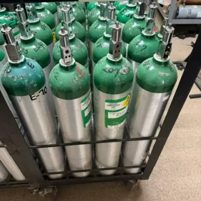 Collection of green and silver aluminum medical oxygen tanks stored in a rolling metal rack, labeled empty.