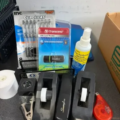 Collection of office supplies including tape dispensers, a Swingline stapler, paper roll, pens, cleaner spray, and a Transcend USB card...