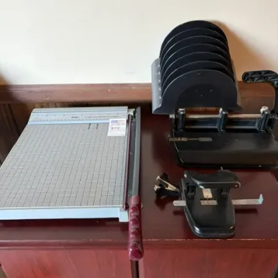 Collection of vintage office supplies including a grid paper cutter, heavy-duty black hole punches, and a staple remover, displayed on a...