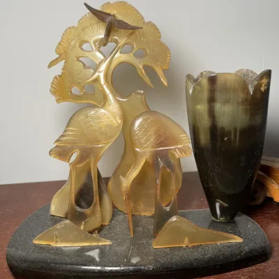 Sale Photo Thumbnail #60: Intricately carved translucent horn sculpture depicting cranes under a pine tree alongside a flower-shaped vase, mounted on a dark stone base.