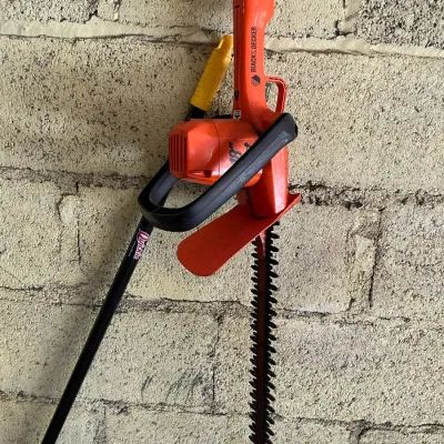 Sale Photo Thumbnail #42: Black & Decker 18-inch corded electric hedge trimmer hanging against a cinder block wall, shown with an adjacent yard tool.