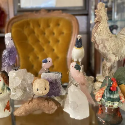 Sale Photo Thumbnail #4: Collection of decorative bird figurines, including hand-carved stone parrots perched on crystal bases, ceramic roosters, and a carved wooden bird.