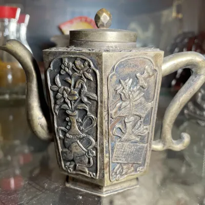 Sale Photo Thumbnail #120: Antique hexagonal metal teapot featuring relief-carved panels with floral designs, displaying an aged patina.