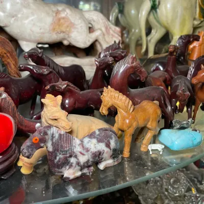 Sale Photo Thumbnail #133: Collection of horse figurines crafted from various materials, including carved wood, polished stone, and ceramics, displayed on a glass shelf.