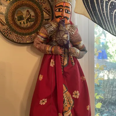 Sale Photo Thumbnail #159: Hand-painted Indian Kathputli string puppet, featuring ornate tinsel detailing and a patterned red skirt, displayed alongside a decorative wall plate.