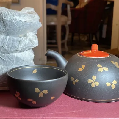 Sale Photo Thumbnail #84: Black ceramic Japanese tea set featuring gold cherry blossom motifs, an orange lid teapot, and matching teacups, partly wrapped in tissue paper.