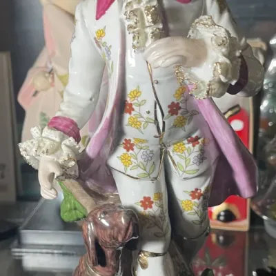 Sale Photo Thumbnail #184: Vintage porcelain figurine depicting an 18th-century nobleman dressed in a pink-lined coat and floral vest, standing beside a small hunting dog.