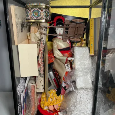 Sale Photo Thumbnail #58: Vintage Japanese doll in traditional kimono displayed inside a black glass case with gold background, surrounded by various books, decorative accessories, and textiles.