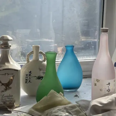 Sale Photo Thumbnail #135: Collection of decorative glassware and ceramic bottles lined up on a windowsill, including frosted glass vases, porcelain decanters, and a Royal Choice Scotch whisky bottle.