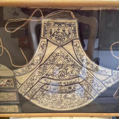 Sale Photo Thumbnail #144: Framed antique Chinese embroidered textile collar or apron, featuring intricate dark blue needlework motifs on off-white fabric, professionally displayed in a wooden shadowbox frame.