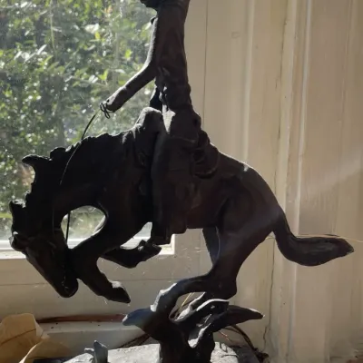 Sale Photo Thumbnail #30: Frederic Remington style bronze statue depicting a cowboy riding a bucking bronco, mounted on a dark stone base.