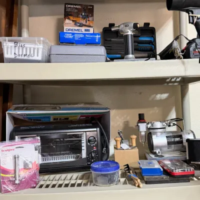 Sale Photo Thumbnail #309: Shelves filled with various crafting and power tools, including a craft/clay oven, Sculpey clay press, Timbertech air compressor, and Dremel accessories.