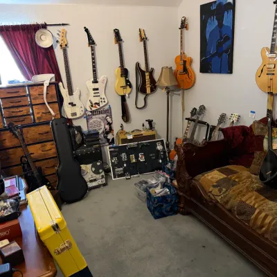 Sale Photo Thumbnail #88: Collection of electric and bass guitars displayed on wall mounts and stands, alongside a wooden daybed sofa, guitar cases, and audio equipment.