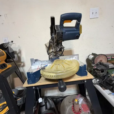 Sale Photo Thumbnail #302: Used blue compound miter saw mounted on a portable stand, shown alongside other power tools and workshop accessories.