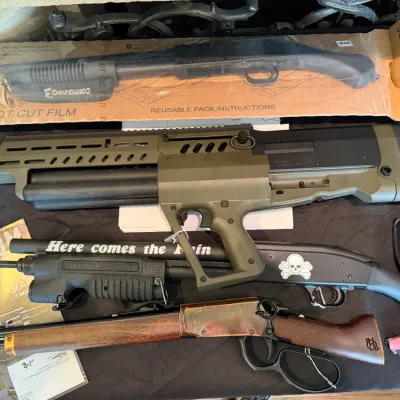 Sale Photo Thumbnail #48: Collection of firearms and accessories, including a bullpup shotgun in olive drab green, a lever-action rifle, a black pistol-grip shotgun, and a pink compact handgun.