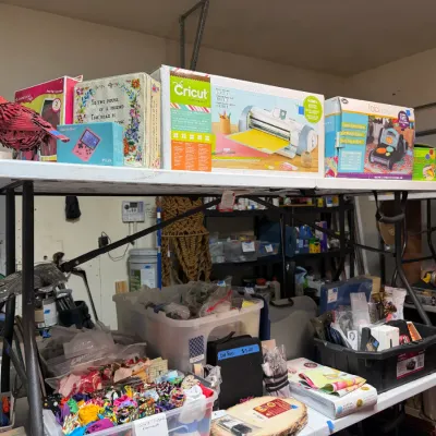 Sale Photo Thumbnail #368: Crafting tools and supplies displayed on folding tables, including a Cricut Expression 2 cutting machine in original box, a Fabi Ellison die cutting machine, and various storage bins filled with fabric scraps and notions.