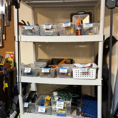 Sale Photo Thumbnail #313: White plastic four-tier shelving unit filled with clear storage bins containing various hand tools, hardware, and extension cords, topped with a novelty N.Y. Yankees Dr. street sign.