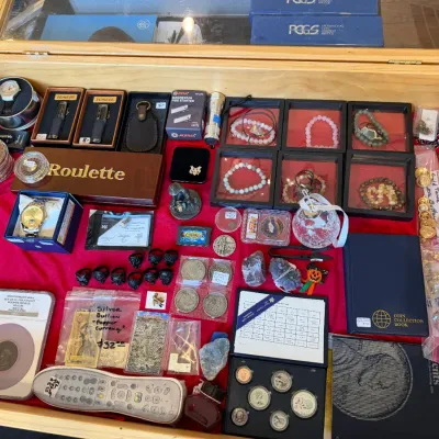 Sale Photo Thumbnail #57: Display case filled with a variety of collectibles including graded coins, silver bullion, beaded bracelets, pocket lighters, coin albums, and small trinkets.